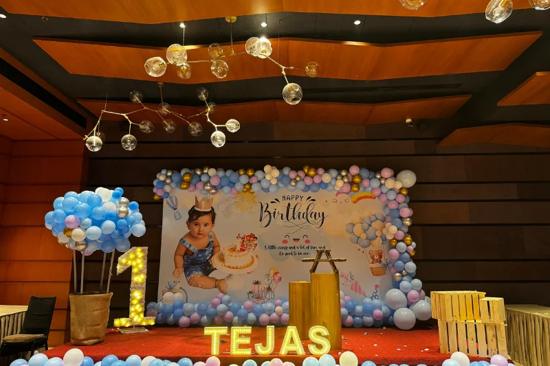 First birthday party stage setup featuring blue and gold balloons, a hot air balloon prop, and illuminated letters spelling Tejas